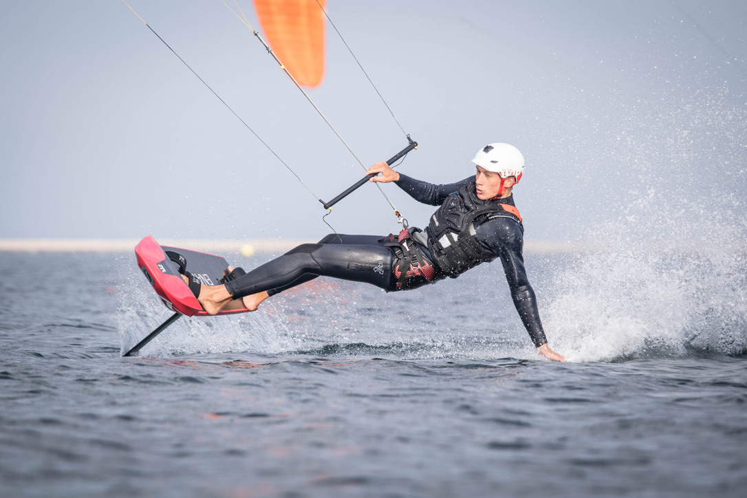 A kitefoiler in helmet and wetsuit leaning hard upwind, spray flying